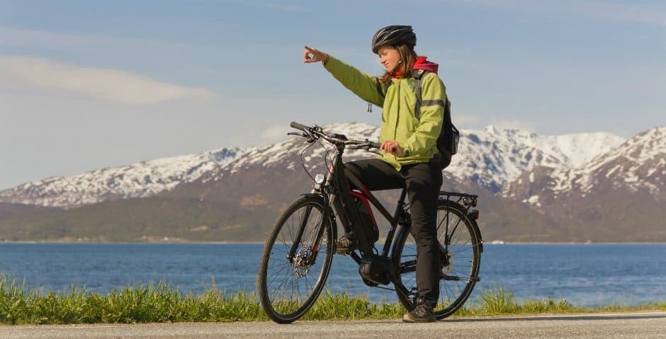 En person sitter på en el-cykel på en väg vid vattnet med ett stort berg i bakgrunden.