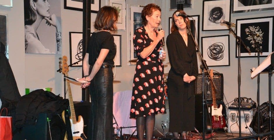 Tre kvinnor står på en scen med gitarrer på och en talar i en mikrofon.