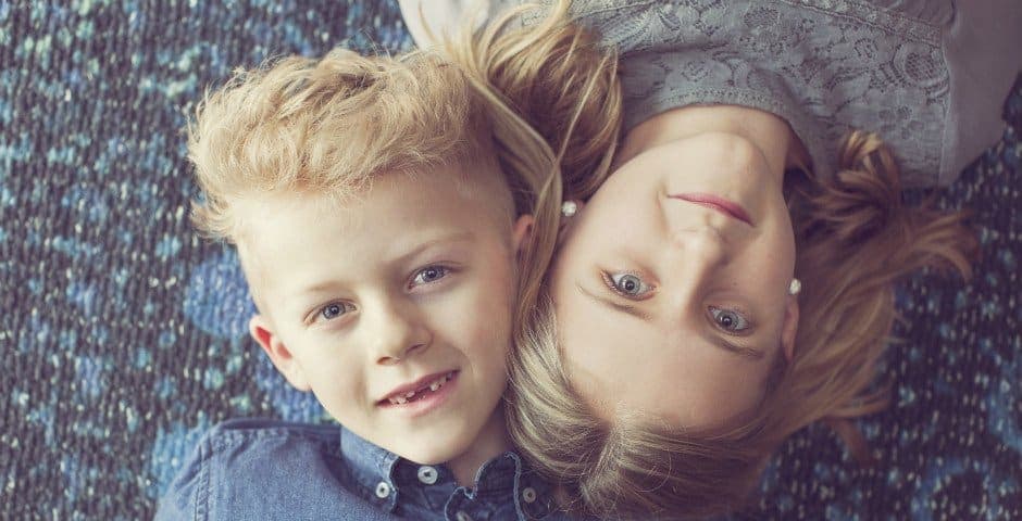 Två barn ligger på golvet med huvuden mot varandra och tittar rakt upp mot kameran.