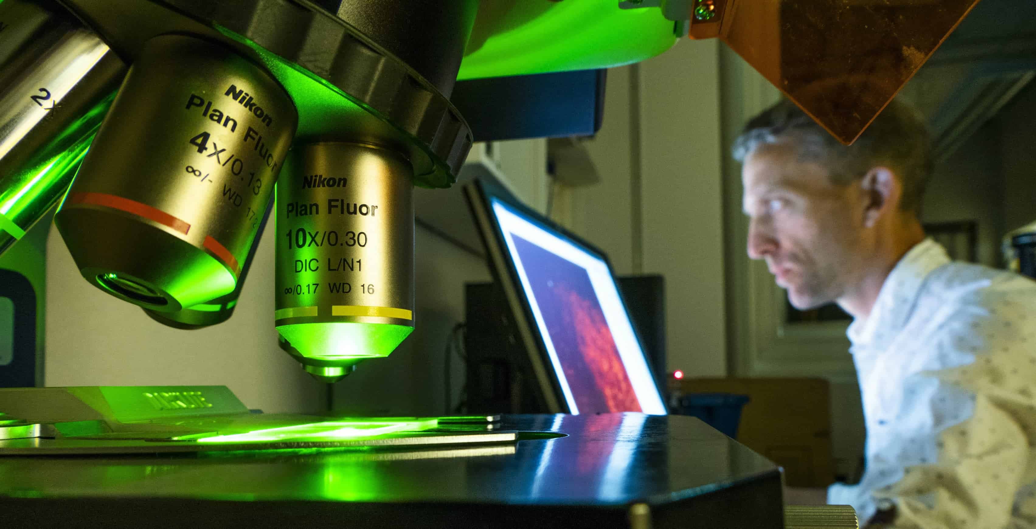 Närbild på ett mikroskop med David Engblom i bakgrunden som sitter vid sin dator.