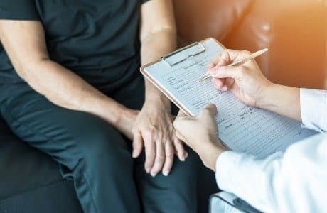 En hand som fyller i ett formulär under ett möte mellan vårdpersonal och en person som sitter på en soffa.