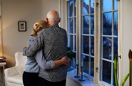 Ett äldre par kramar om varandra i sitt hem vid fönstret.