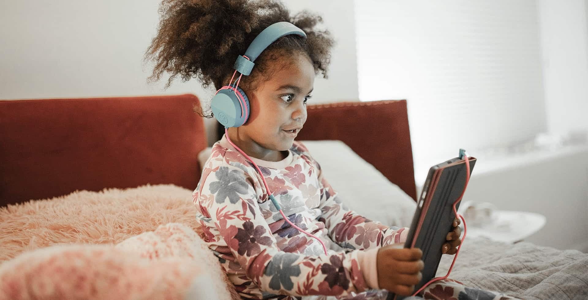 En flicka sitter i en säng med hörlurar och iPad.