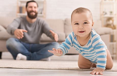 En bebis kryper på ett golv med sin pappa bakom sig.
