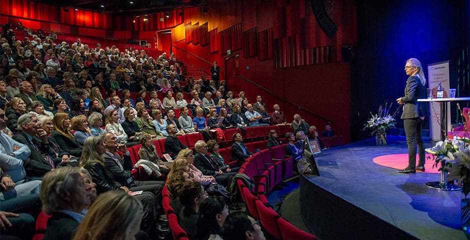 Publik i en sal som tittar mot en scen där en person står och talar.