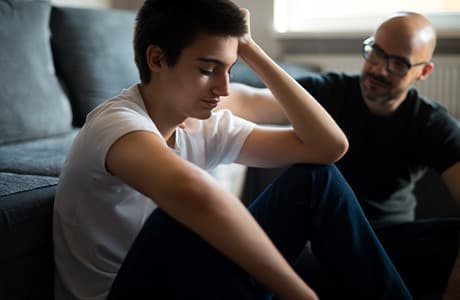 Ung kille sitter på golvet vid en soffa och hans pappa sitter bredvid med handen på axeln.