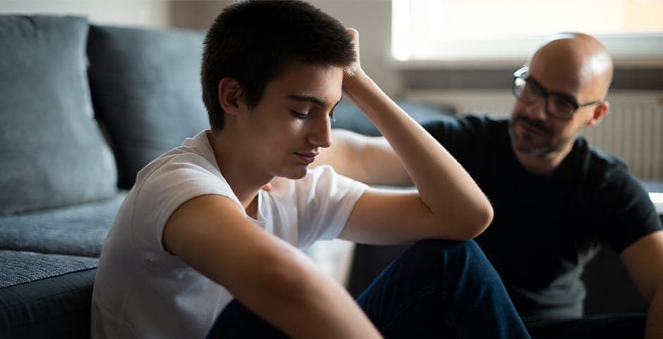 Ung kille sitter på golvet vid en soffa och hans pappa sitter bredvid med handen på axeln.