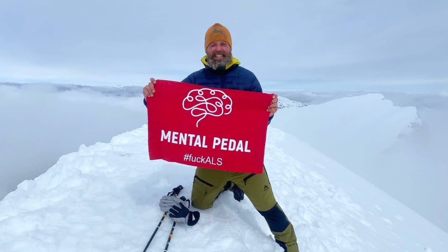 Fredrik Lundgren står på toppen av Kebnekaise med en flagga där det står "Mental Pedal. #fuckALS".