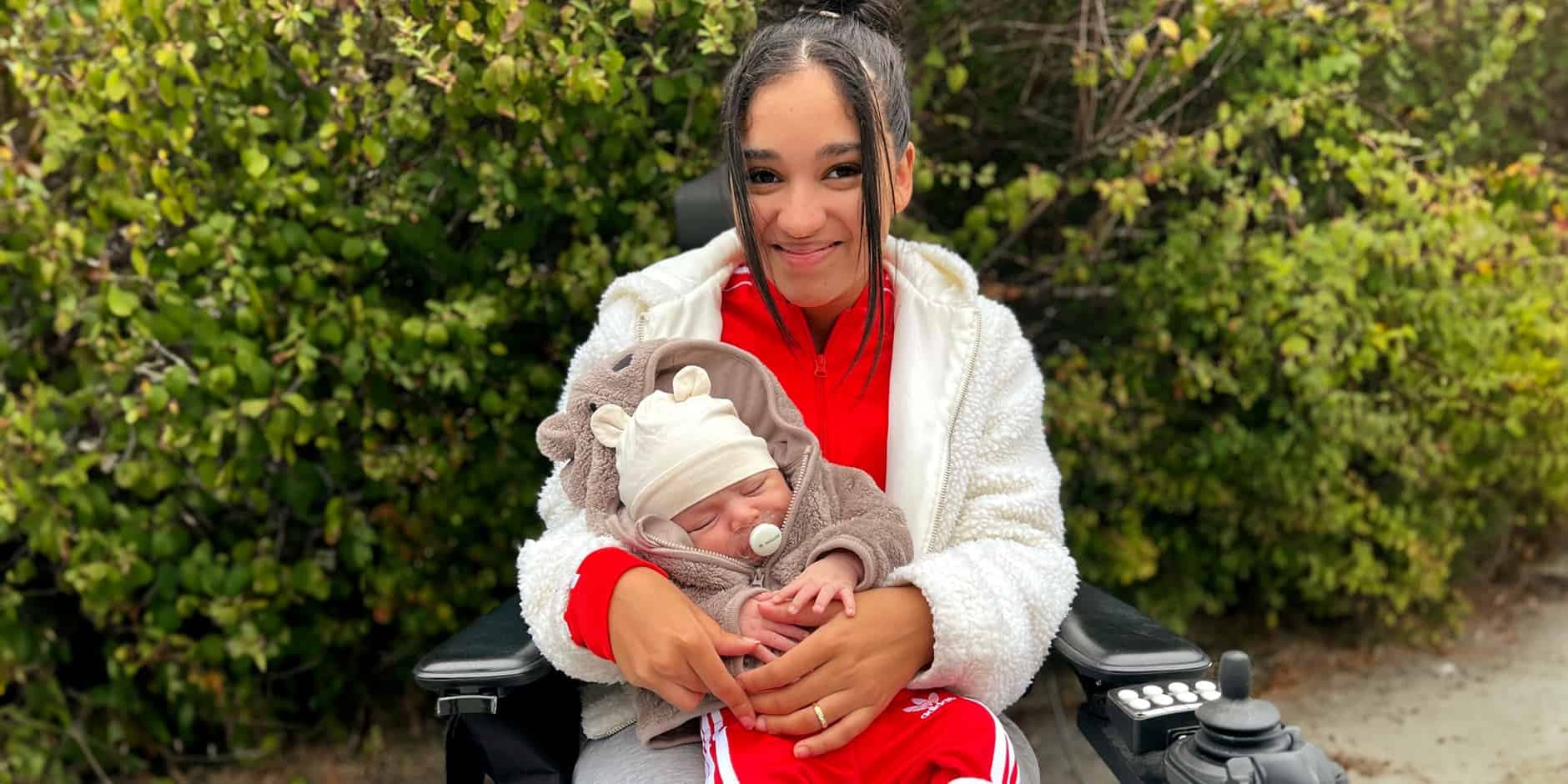 Carolina Hamnefjäll sitter framför ett buskage i sin rullstol med sin son Matheus i famnen.