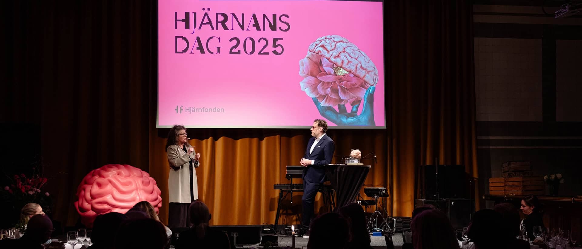 Två talare på en scen framför publik och en skärm duk där det står Hjärnans dag 2025.
